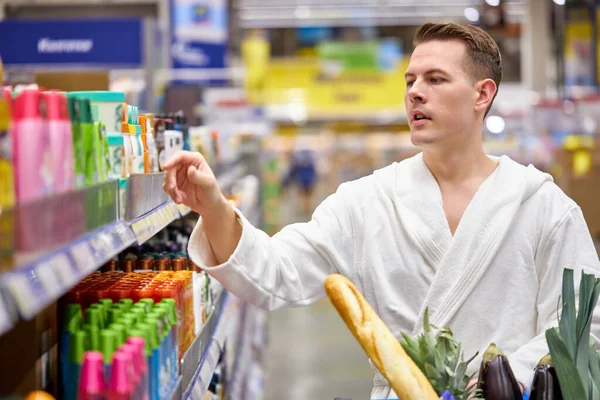 Young good-looking man need new shampoo or shower gel, came in supermarket in bathrobe