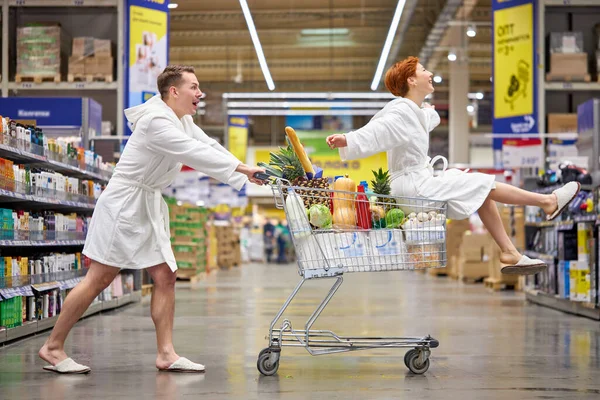 Вид сбоку на возбужденную веселую пару в белых халатах, весело катающуюся по магазинам и сидящую на ней — стоковое фото