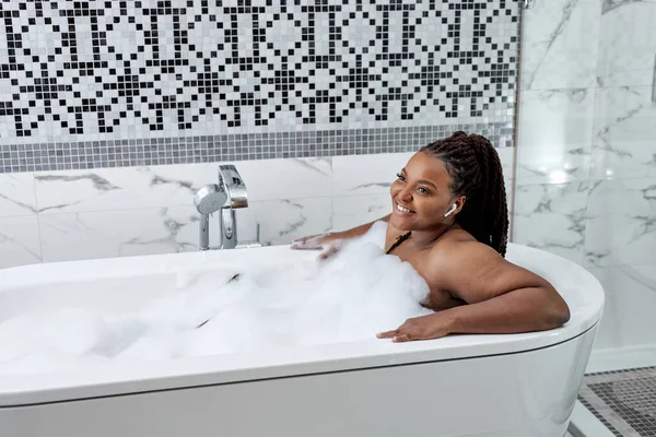 Relaxante na banheira. Jovem mulher africana se divertindo no banheiro, nos fones de ouvido — Fotografia de Stock