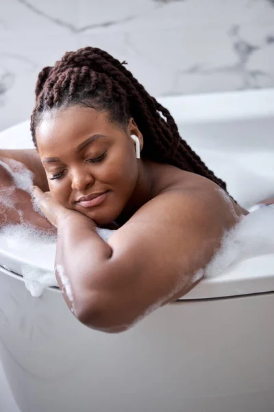Calma dama afroamericana soñadora acostada en la bañera con espuma, relajante en los auriculares —  Fotos de Stock
