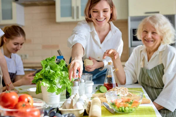 Χαρούμενη Καυκάσια ενήλικη γυναίκα που παίρνει αλάτι ενώ μαγειρεύει, με την οικογένεια στην κουζίνα — Φωτογραφία Αρχείου