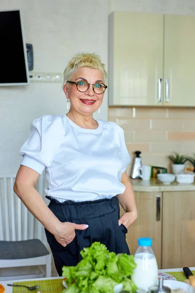 elderly caucasian woman in eyeglasses posing at camera in kitchen at home, smiling