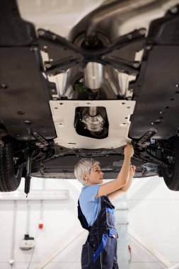 Garajda, arabanın altında çalışan beyaz kadın oto tamircisi. Kaldırılan araba denetleniyor