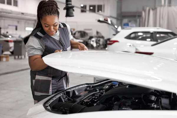 Jonge Afrikaanse vrouw in uniform is het openen van de kap van de witte auto in auto reparatiewerkplaats — Stockfoto