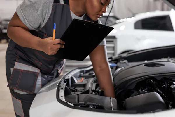 Afrikaanse vrouwelijke monteur op het werk, controleren auto kap schrijven notities in tablet — Stockfoto