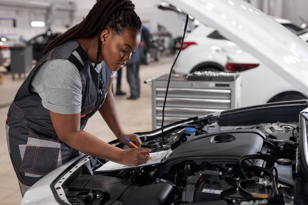 Afrikaanse vrouwelijke monteur op het werk, controleren auto kap schrijven notities in tablet — Stockfoto