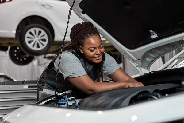 Smiling african lady in overall repairing auto hood, using instruments tools