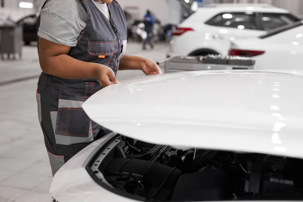 Bijgesneden zwarte vrouw in uniform is het openen van de kap van de witte auto in auto reparatiewerkplaats — Stockfoto