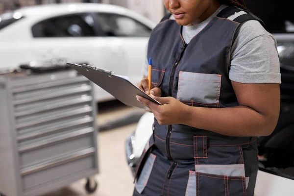 Zwart vrouwelijk auto monteur maken auto Checkup, in uniform schrijven en houden Klembord — Stockfoto