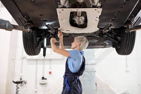 Zijaanzicht op vrouwelijke auto monteur werken onder auto in de garage. inspectie van opgeheven auto — Stockfoto