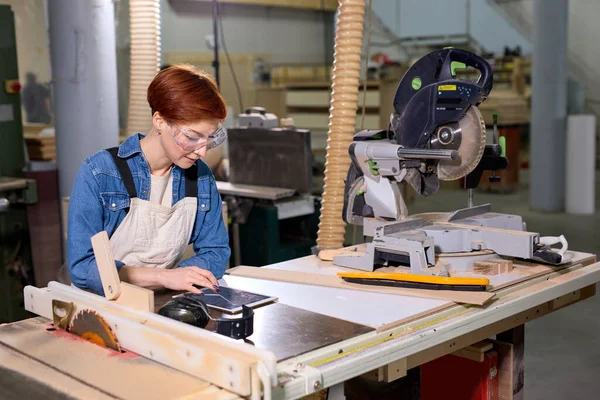 Vista lateral no marceneiro feminino confiante usando tablet digital enquanto trabalhava na oficina de carpintaria — Fotografia de Stock