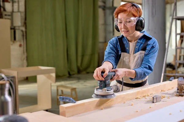 Jovem carpinteiro feminino trabalhando com madeira usando lixadeira elétrica em oficina — Fotografia de Stock