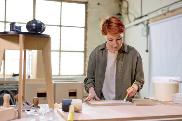 Mulher carpinteiro artesanal pintura com escova e pintar a tábua de madeira na oficina — Fotografia de Stock