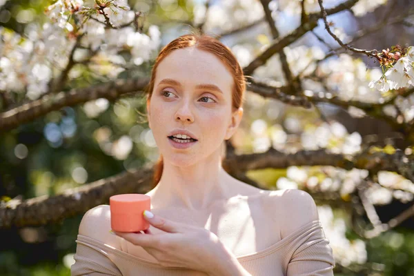 Fêmea de boa aparência usando creme hidratante posando no jardim da primavera com árvores florescendo — Fotografia de Stock