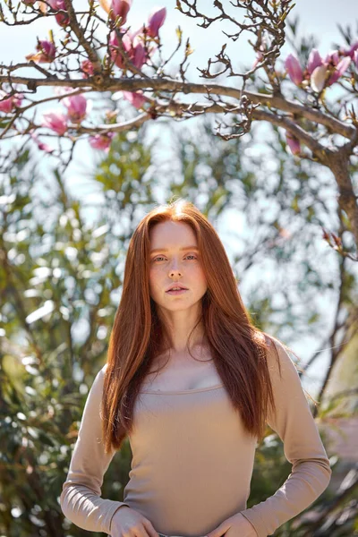 Dame met lang natuurlijk rood haar poseren aan de camera tegen de achtergrond van de boom met roze bloemen — Stockfoto