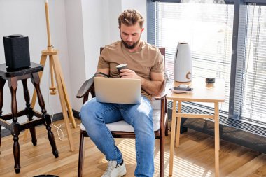 Yakışıklı genç adam evde bilgisayar kullanıyor. Rahat rahat ev içi aktiviteler yapıyor.