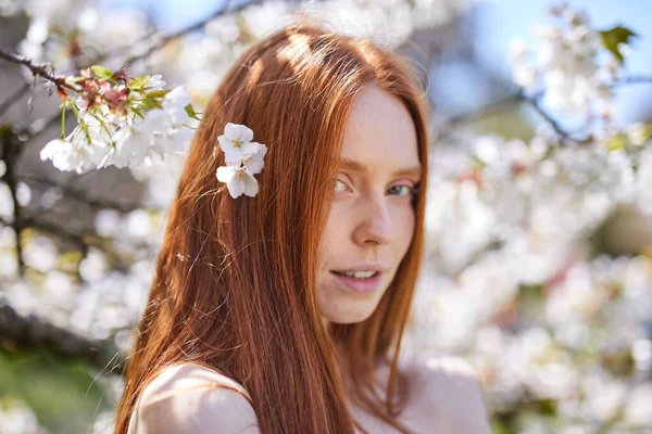 Kızıl saçlı güzel kadın çiçek açan ağaçların tadını çıkarıyor. Bahar Bahçesinde Moda Modeli. — Stok fotoğraf