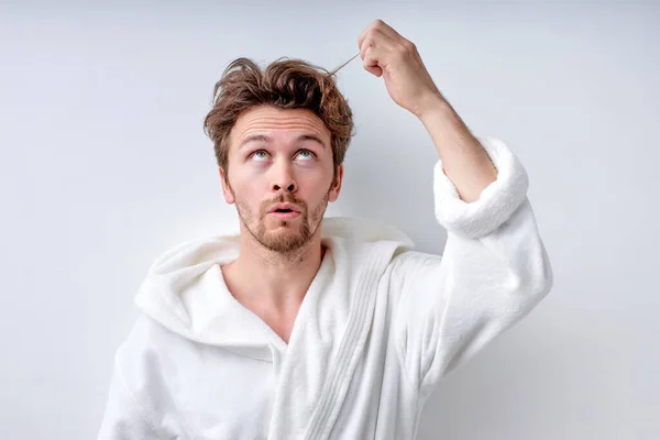 Shaggy confuso ragazzo in accappatoio isolato su sfondo bianco, tocca lunghi capelli ricresciti — Foto Stock