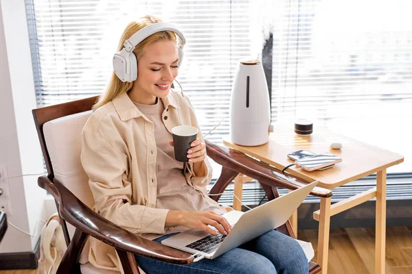 Mujer rubia caucásica se sienta a trabajar en el ordenador portátil, en los auriculares, el uso de humidificador —  Fotos de Stock