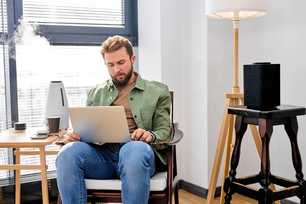 Guy arbeiten am Laptop im Zimmer mit tragbarem Lautsprecher, Luftreiniger, im Smart House allein — Stockfoto