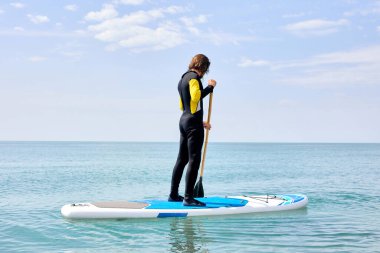 Siyah dalgıç kıyafetli genç adam sörf tahtasında kürekle okyanusta süzülüyor.