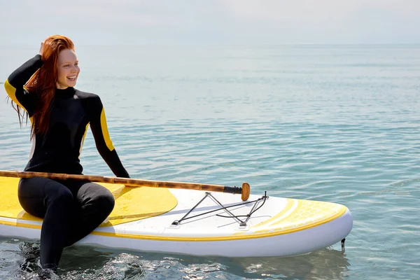 Rothaarige schöne Frau paddelt am Strand, sitzt auf dem Paddelbrett, erholt euch — Stockfoto