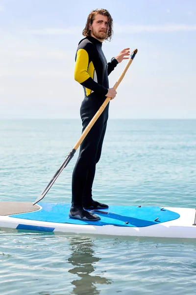 Junger Mann im schwarzen Neoprenanzug mit Paddel auf Unterbord schwimmt auf dem Wasser im Ozean — Stockfoto