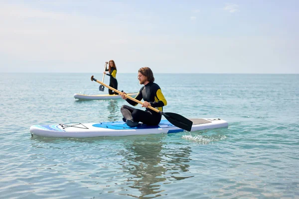 Vista lateral em casal levante-se paddleboarding no mar. Jovem casal fazendo watersport — Fotografia de Stock