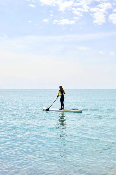 Rückansicht einer Frau im schwarzen Neoprenanzug mit Paddel auf dem Unterbord schwimmt auf dem Wasser im Ozean — Stockfoto