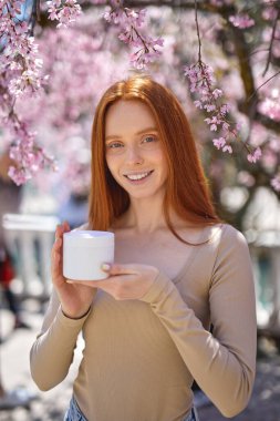 Mutlu gülümseyen, doğal makyajlı güzel bir kadın. Krema kavanozuyla poz veriyor. Çiçeklerin yanında.