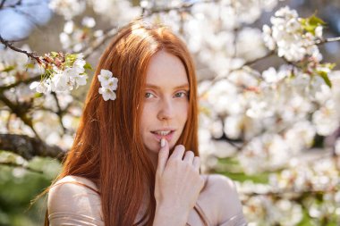 Kızıl saçlı güzel kadın çiçek açan ağaçların tadını çıkarıyor. Bahar Bahçesinde Moda Modeli.