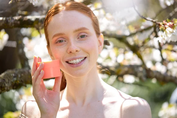 Open-minded caucasian lady with natural make-up holding cream jar in hands