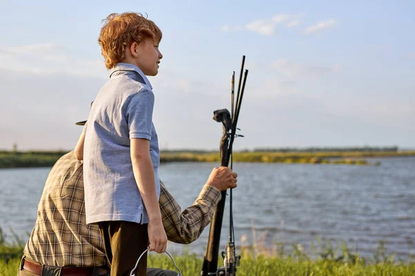 Adorable sweet redhead teenage boy came with granddad to fish, looking at side