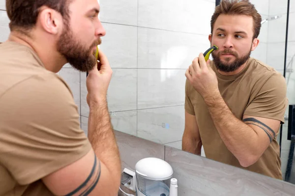 Ομορφιά, ξύρισμα. άντρας κοιτάζεται στον καθρέφτη, ξυρισμένη γενειάδα με ηλεκτρικό ξυριστικό — Φωτογραφία Αρχείου