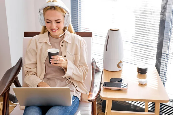 Blonde Kaukasierin sitzt am Laptop, in Kopfhörern und benutzt Luftbefeuchter — Stockfoto