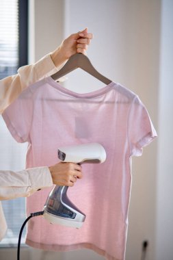 Evdeki sıradan tişörtleri ütülemek için buharlı ütü kullanan tanınmamış bir kadın.