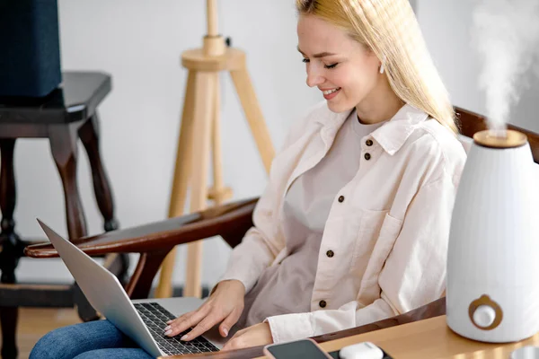 Hübsche blonde Frau arbeitet an Laptop in der Nähe von Luftreiniger und Feuchtigkeitscreme-Gerät — Stockfoto