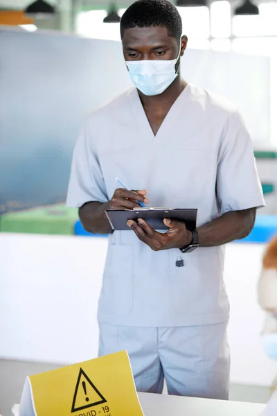 Beyaz üniformalı Afro-Amerikan tıp çalışanı hastanede pano yazısıyla ayakta duruyor. — Stok fotoğraf