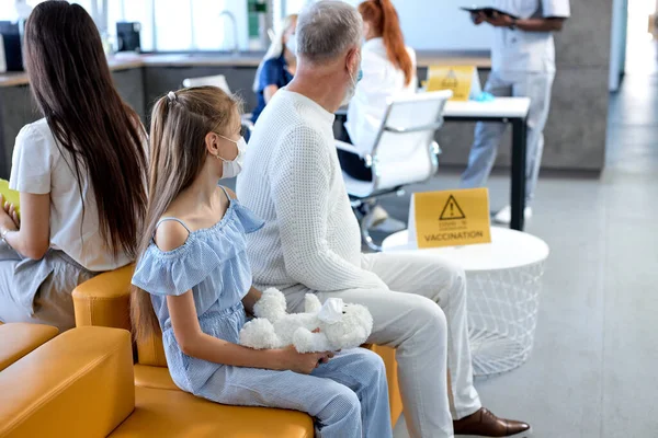 Niña con juguete se sienta en el hospital esperando la vacunación, mirando al costado —  Fotos de Stock