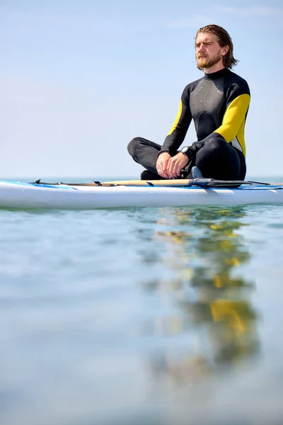 Pensativo deportista tranquilo en traje de neopreno sentado en la tabla de surf esperando una gran ola —  Fotos de Stock