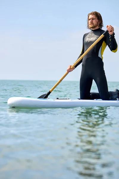 Homem de fato de mergulho em pé na prancha de remo no mar em água azul. Modelo masculino vista lateral — Fotografia de Stock