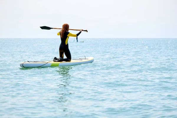 Visão traseira sobre a mulher em wetsuit levantando remo enquanto navega, desfrutar de estilo de vida ativo praia — Fotografia de Stock