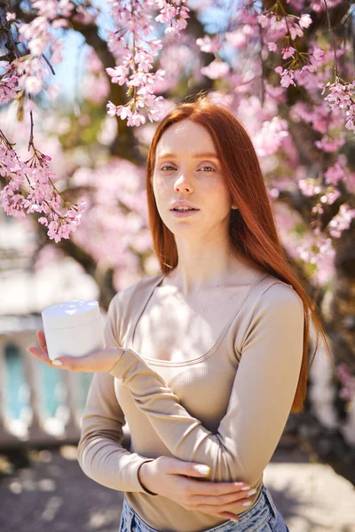 Calm beautiful female with natural makeup posing with cream jar, near blooming flowers