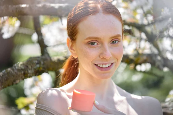 Open-minded caucasian lady with natural make-up holding cream jar in hands