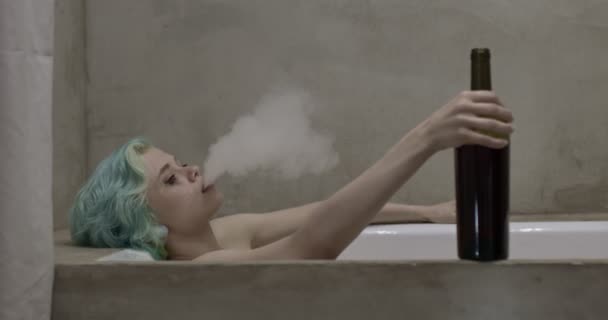 Долористая молодая женщина с зелеными короткими волосами, лежащими в ванне, с бутылкой вина — стоковое видео