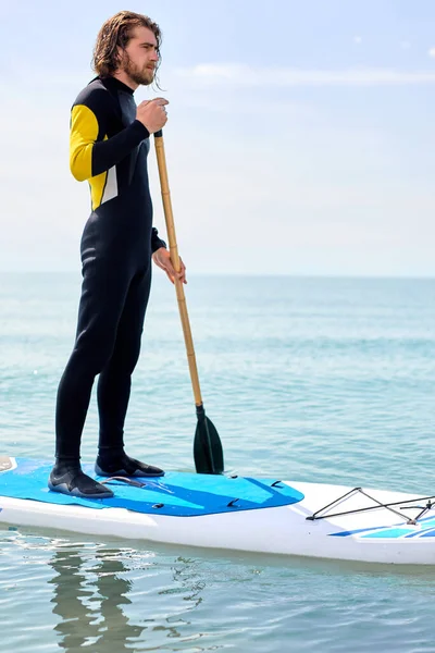 Man with long hair in black wetsuit working out with stand up paddle board on water — Fotografia de Stock