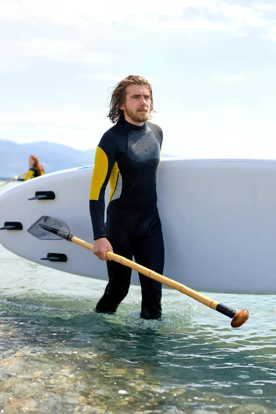 Macho joven en traje de buceo spedting tiempo en el mar, llevando tabla de surf y remo —  Fotos de Stock