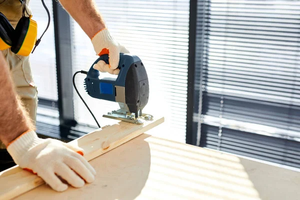 Cropped construction worker cuts wooden board with an electric jigsaw while renovating house — Fotografia de Stock