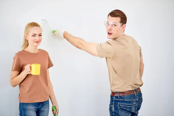 Funny caucasian married couple paint the wall, woman have rest, while husband is painting — Fotografia de Stock