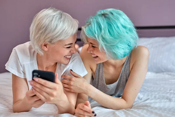 Twee jonge aantrekkelijke vrouwen thuis liggen op bed met smartphone, plezier hebben — Stockfoto